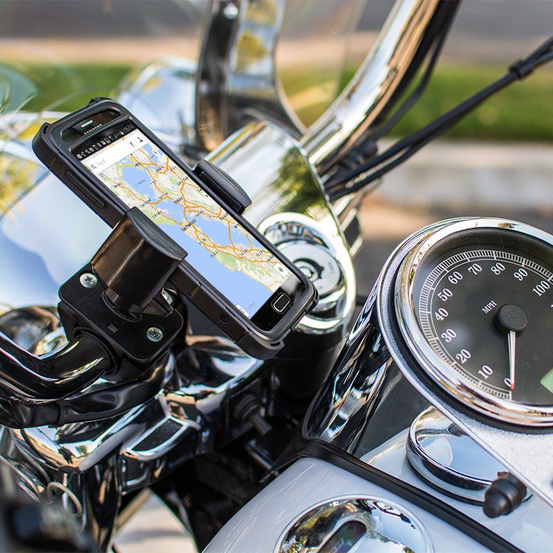 A RoadVise® Holder with Handlebar Mount secures a smartphone to motorcycle handlebars beside the speedometer, with blurred greenery and a street visible in the background.