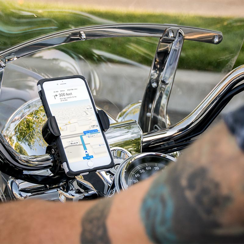 A smartphone with a GPS map is secured on motorcycle handlebars using the RoadVise® Motorcycle Phone Mount - Black Aluminum. A tattooed arm appears in the foreground, while chrome parts of the motorcycle reflect the light.