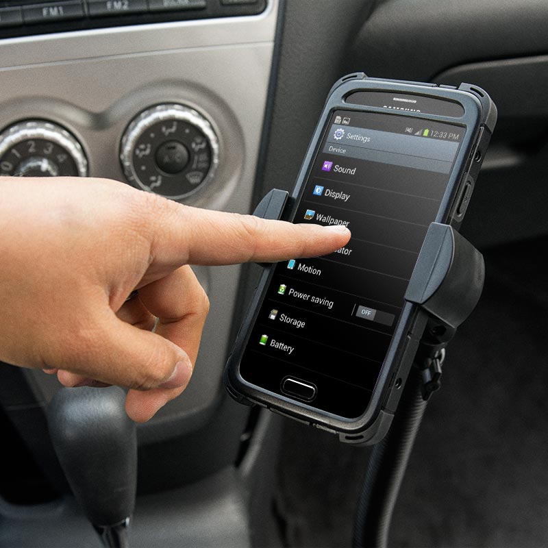 A hand touches a smartphone displaying the settings menu, mounted on the RoadVise® Phone Holder with 15 Gooseneck and Seat Rail Mount in a car, with car controls and gear shift visible in the background.