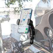 A RoadVise® Phone Holder with Aluminum Handlebar Mount and 2.75 Shaft secures a smartphone displaying a navigation map on a motorcycle handlebar, with an outdoor background featuring a blurred wall and trees.