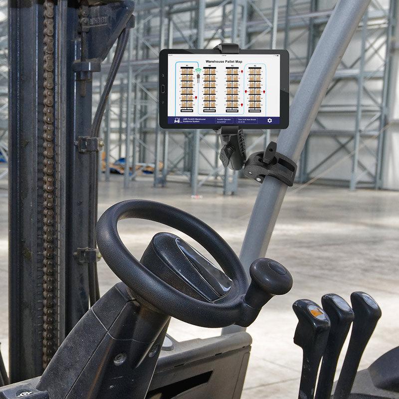A forklift cab has a RoadVise® Ultra Phone and Tablet Holder with Clamp Mount and Security Knob securing a tablet that displays a warehouse pallet map. The steering wheel, control levers, and an organized warehouse are visible in the background.