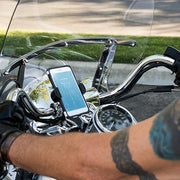 A close-up of a gloved hand gripping chrome handlebars shows the RoadVise® XL Motorcycle Midsize Tablet and Phone Mount—Chrome Aluminum—displaying a weather app at 78°F. Chrome accents and a tattooed arm are partially visible.