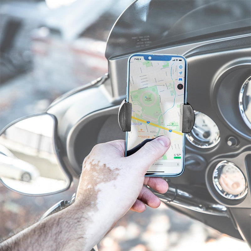 A hand with vitiligo holds a smartphone secured in the RoadVise® XL Phone and Midsize Tablet Holder with Handlebar Collar Mount and 2.75 Shaft on a motorcycle dashboard, displaying a map app above the bike’s handlebars and gauges.