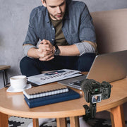 A man sits at a table with a laptop, notepad, and BUSINESS magazine. A camera attached to a Robust Clamp Camera Mount with Security Knob on a tripod records him; his focused image is visible on the screen as he looks down.