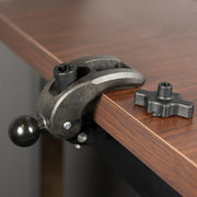 The Robust Clamp Mount with Security Knob - 25mm (1 inch) Compatible is attached to the edge of a wooden table, with a separate cross-shaped black plastic piece and a 25mm ball end placed next to it.