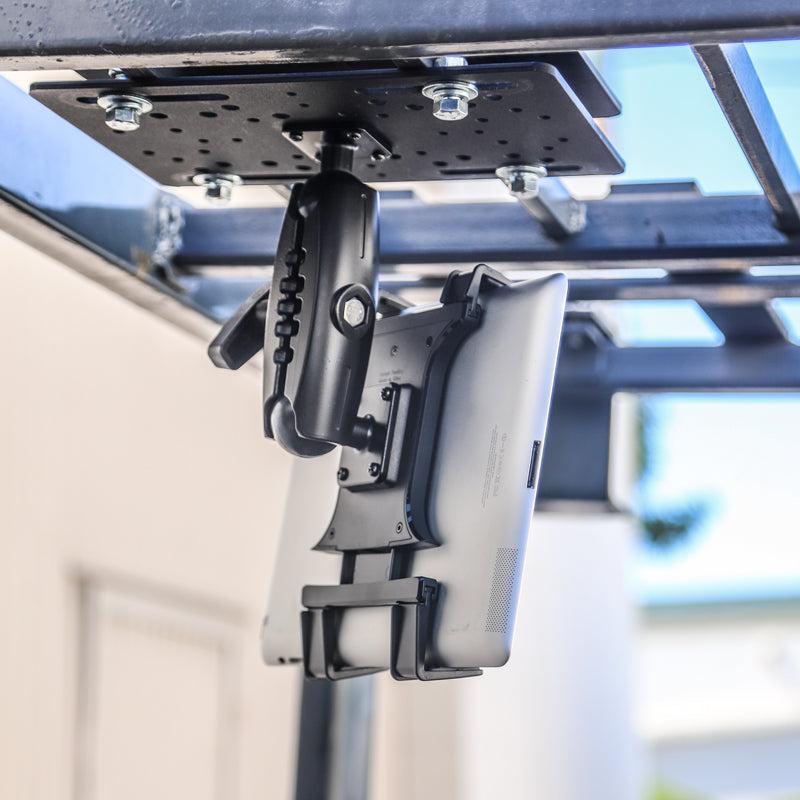The Slim-Grip® Overhead Guard Forklift Tablet Mount with 5 Shaft is securely installed upside down on a metal frame outdoors near a building wall using its adjustable bracket and locking mechanism.