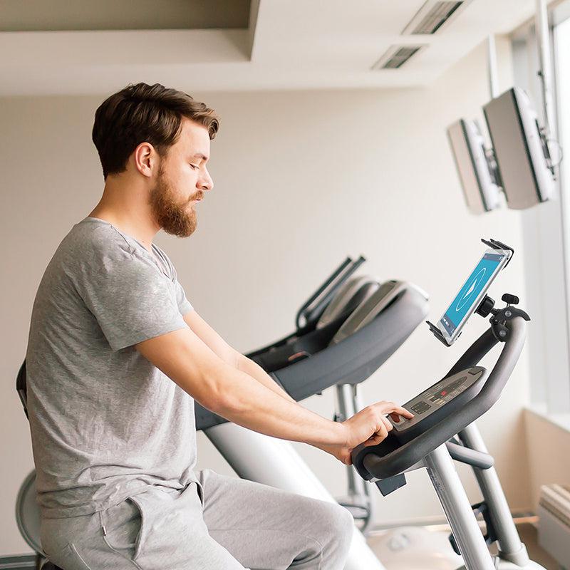 A man in grey workout clothes uses a stationary bike in a gym, viewing a screen secured by the Slim-Grip® Tablet Clamp Mount attached to the handlebars. Other gym equipment and large windows can be seen in the background.