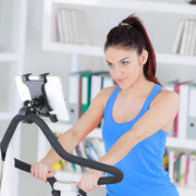 A woman in a blue tank top rides indoors on a stationary bike, using the Slim-Grip® Tablet Clamp Mount for Spin Bike attached to the handlebars to view her device. Bookshelves with books and folders appear in the background.