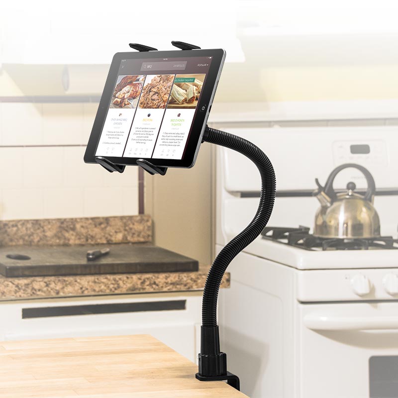 The Slim-Grip® Tablet Holder with Clamp Mount and 22 Gooseneck is clamped to the kitchen counter, securely displaying recipes on a tablet, with a stove, kettle, and other appliances visible in the background.