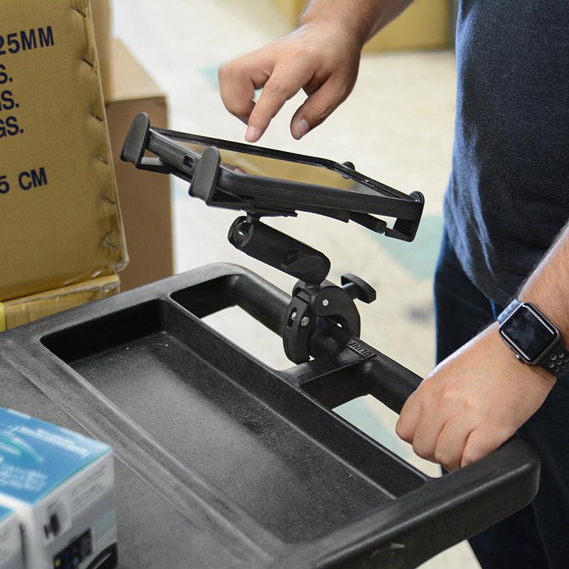 A person adjusts the Slim-Grip® Tablet Holder with Clamp Mount and Security Knob Shaft on a cart handle while wearing a smartwatch, with boxes stacked in the background.
