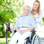 An elderly woman in a wheelchair smiles outdoors as a young woman stands behind her. The Slim-Grip® Tablet Holder with Height-Adjustable Desk Stand is positioned beside the wheelchair, offering easy tablet access in the lush, green setting.