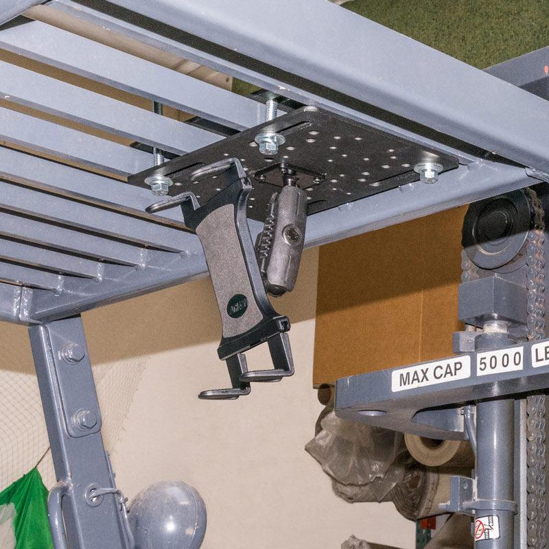 Close-up of a Slim-Grip® Tablet Holder with Overhead Forklift Guard Mount and 3.75 Shaft attached under a metal rack labeled MAX CAP 5000 LB, with various objects and storage items visible in the background.
