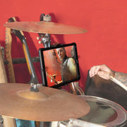 A person plays drums in a red-walled room with guitars behind. An iPad, held by a Slim-Grip® Tablet Mount with Clamp Base on the drum set, shows his focused face as he performs.