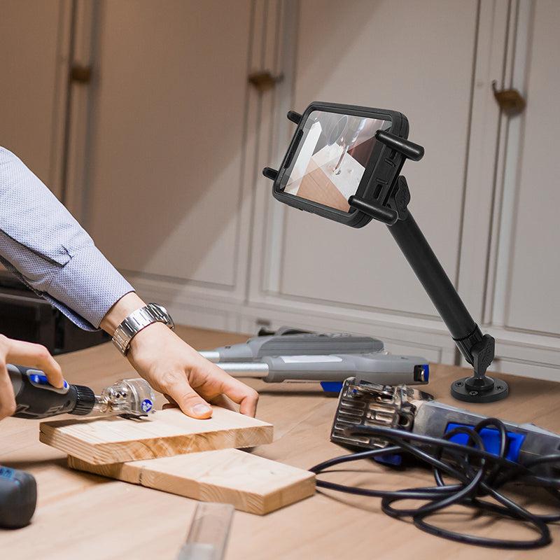 A person carves wood on a workbench, with tools nearby, while a Slim-Grip® Ultra Heavy-Duty Drilled-Base Midsize Tablet Mount holds a tablet facing the workspace to record or display the activity.