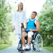 A smiling man in a wheelchair with a bandaged knee and a woman in a white coat sit outdoors, live streaming together on the Slim-Grip® Ultra iPad mini Desk Stand Tablet Holder (17–29” Tall), surrounded by greenery.