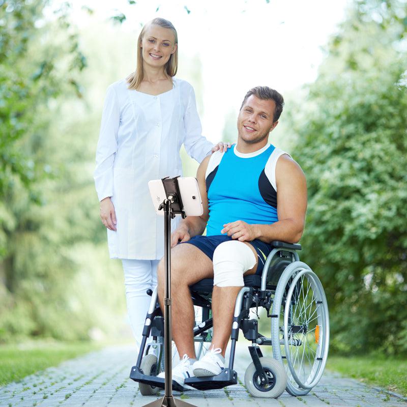 A smiling man in a wheelchair with a bandaged knee and a woman in a white coat sit outdoors, live streaming together on the Slim-Grip® Ultra iPad mini Desk Stand Tablet Holder (17–29” Tall), surrounded by greenery.