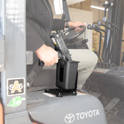 A person in a black jacket and tan pants places a handheld barcode scanner into the SteadyMag™ Magnetic Mount System with Large Barcode Scanner Holder, mounted beside the seat of a Toyota forklift, with controls and part of the cab visible.