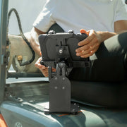 A person in a white shirt holds a rugged, black tablet secured by the SteadyMag™ Magnetic Mount System with LockVise™ Locking Tablet Holder, seated on industrial equipment.