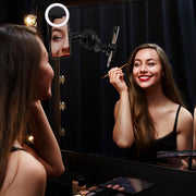 A woman in a black dress smiles as she applies makeup before a mirror, using the Suction RoadVise® Phone Mount and Ring Light Bundle to illuminate her beauty setup. Makeup products are arranged on the counter beneath the gentle ring light glow.