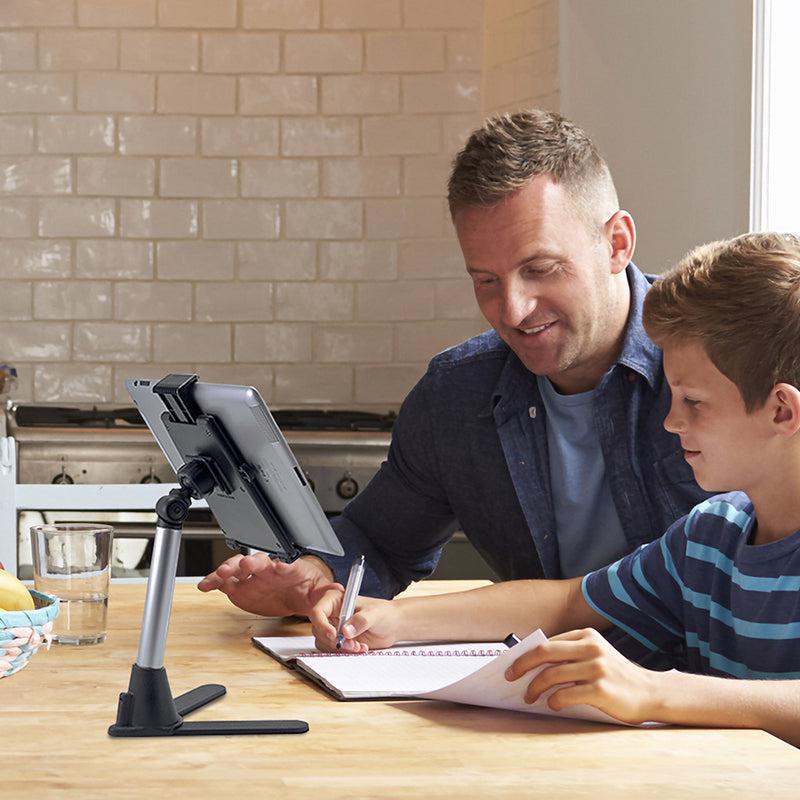 A man and a boy sit at a kitchen table, using a Desk Stand Mount with Tablet Holder. The boy writes in a notebook while the man smiles supportively. Behind them are kitchen appliances and a tiled wall.