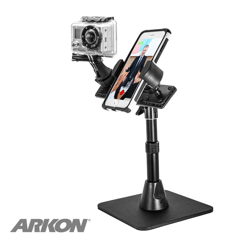 A black TW Broadcaster Desk Stand for GoPro and RoadVise® Phone Holder displays a smartphone with a person in formal attire and a GoPro camera. Adjustable arms and a flat base make it ideal for live streaming. The Arkon logo is in the bottom left corner.