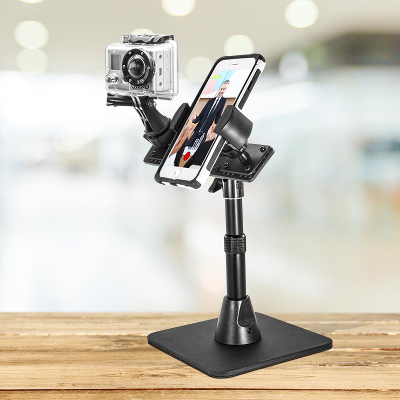 The TW Broadcaster Desk Stand for GoPro and RoadVise® Phone Holder is mounted side by side on a wooden table, perfect for content creators live streaming or filming videos with a blurred indoor background.