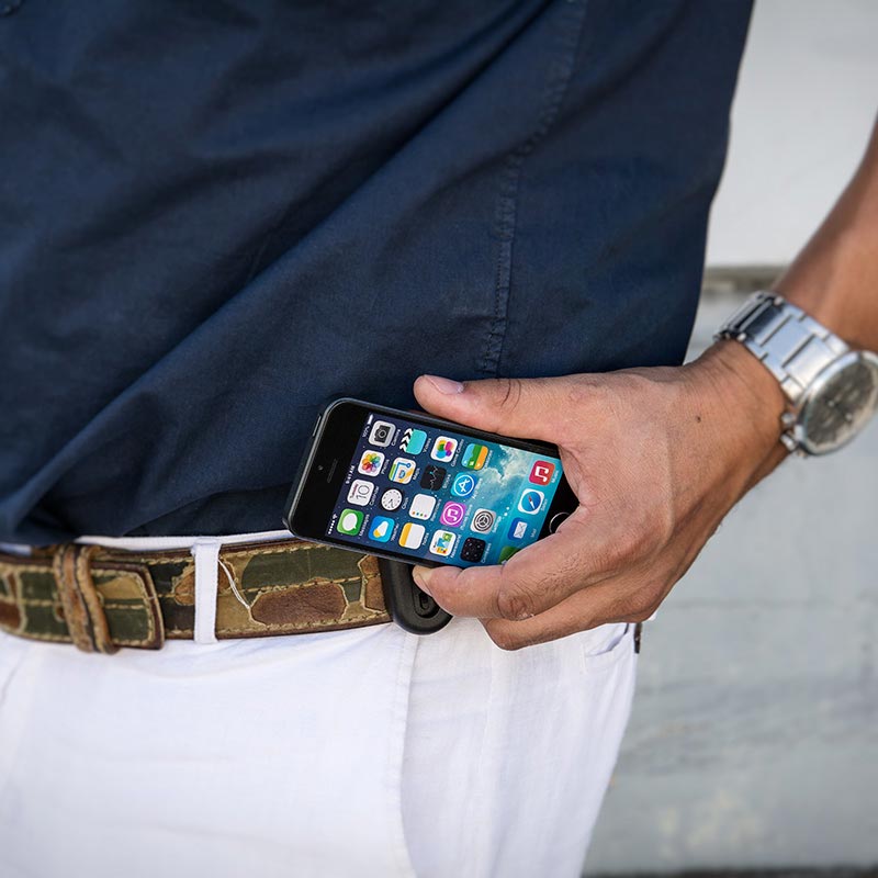 A person in a navy shirt and white pants wears a camo Universal Belt Clip Holder for Smartphones, with their phone showing apps. They also have on a silver wristwatch.